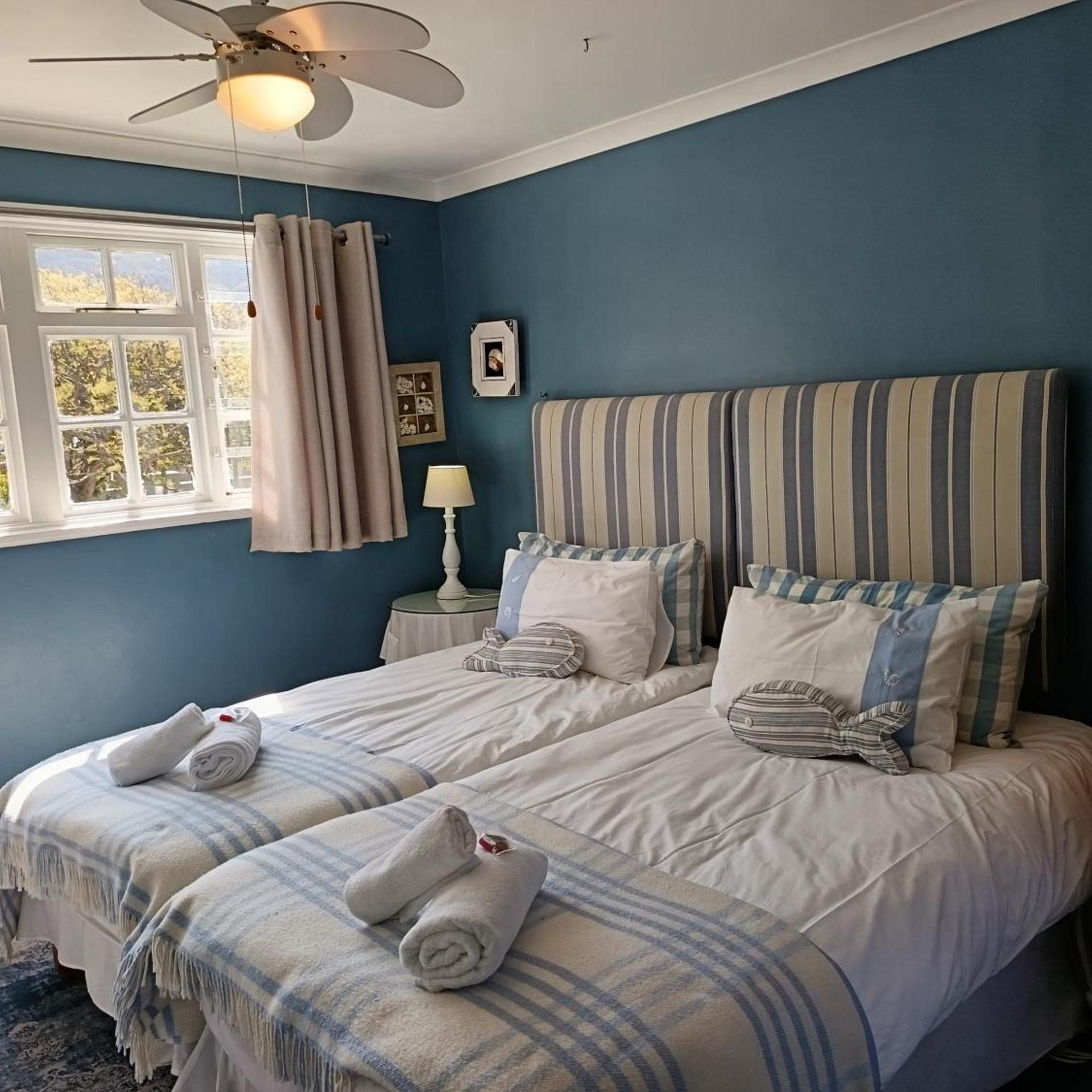 A bedroom with two beds, a ceiling fan, a window with curtains, and a bedside table with a lamp.