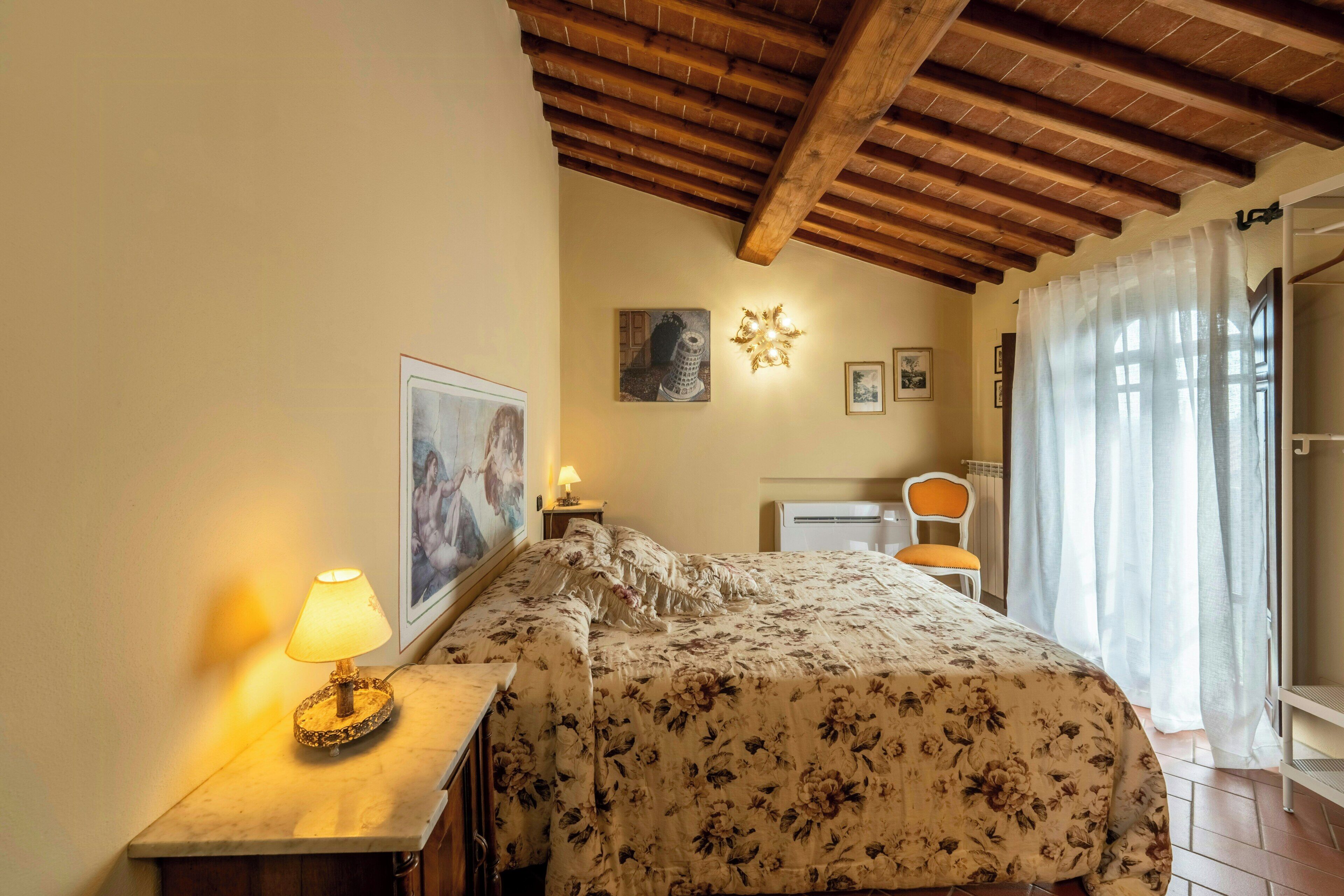 Ein Schlafzimmer mit einem Bett, einem Nachttisch, einer Lampe, einem Gemälde, einer Wanddekoration, einem Sessel und einem Fenster mit Vorhängen.