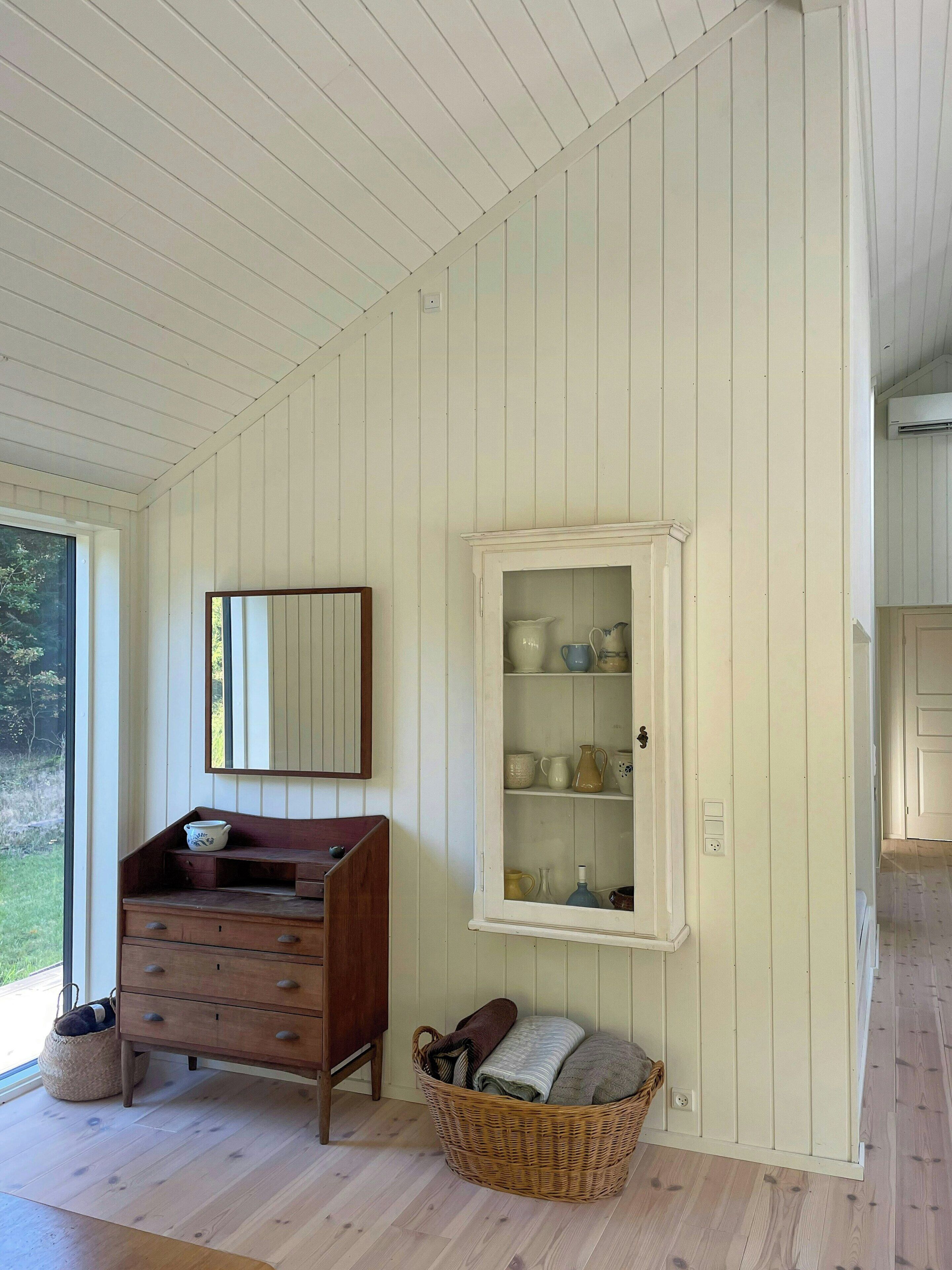 A room with a wooden dresser, a mirror, a glass cabinet with pottery, and a wicker basket with towels.