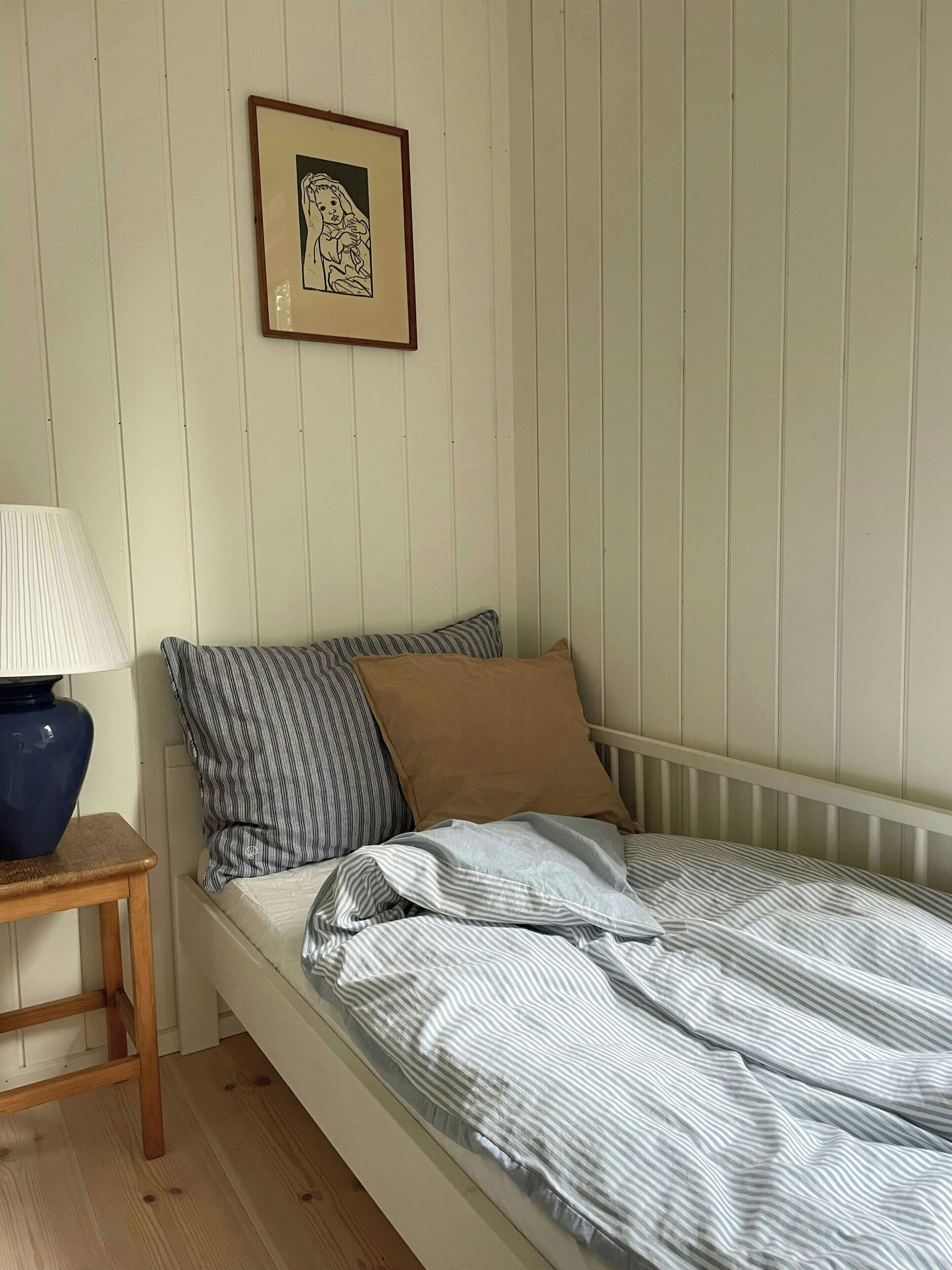 Un lit simple avec une literie à rayures, un oreiller marron et un autre à rayures, le tout placé devant un mur blanc orné d’un tableau encadré.