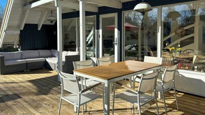 Une terrasse extérieure avec une table et des chaises en bois, un canapé blanc et une porte vitrée donnant sur l’intérieur.