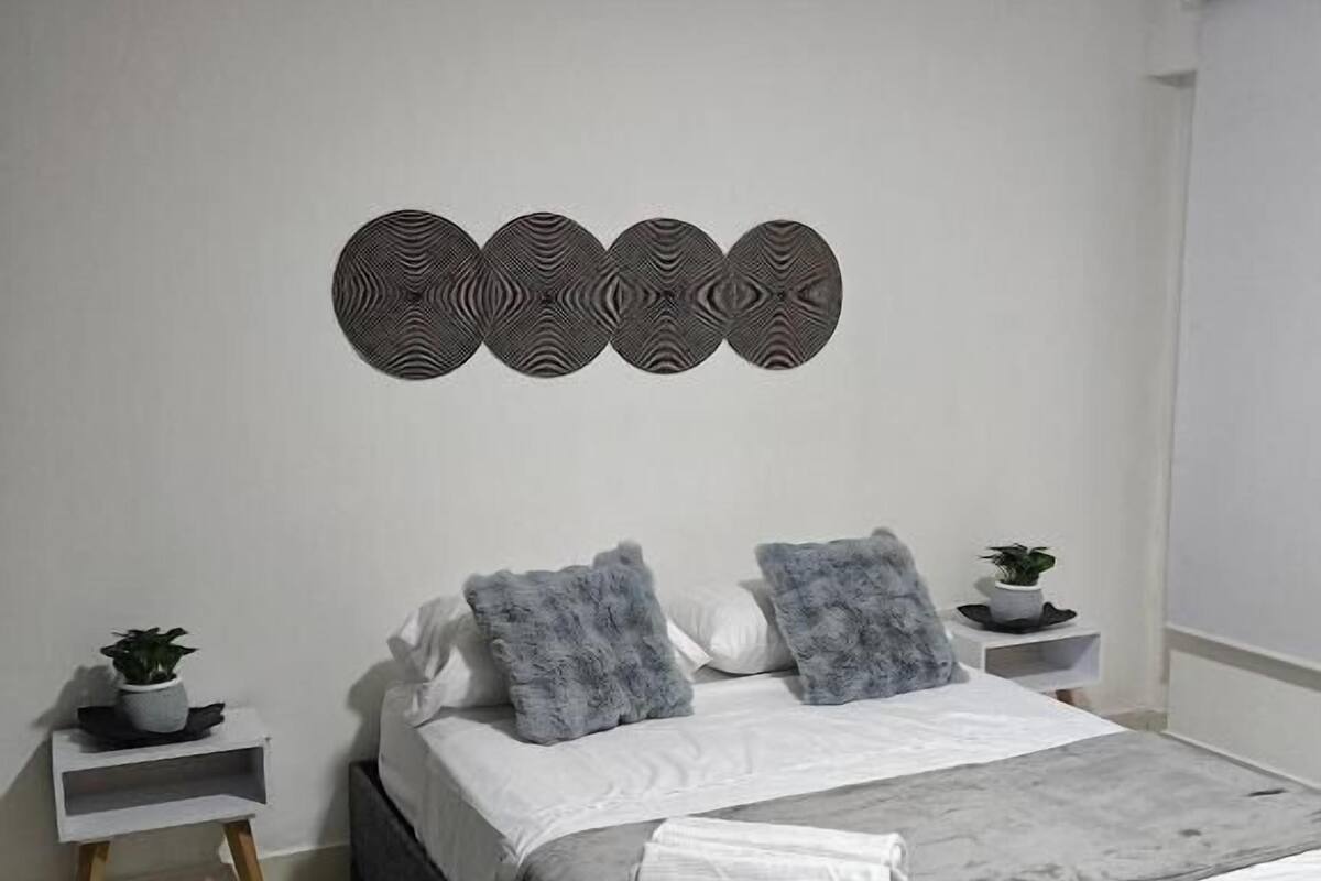 A modern bedroom with a grey bed, white bedding, two grey pillows, a grey throw blanket, a wall-mounted circular decoration, and a small plant on a bedside table.