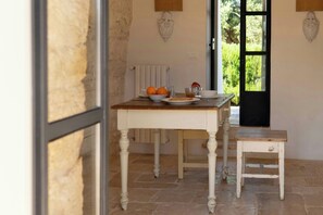 A dining table with a bowl of oranges and a plate, a small stool, and a window with a view of greenery. - pool villa among ancient olive trees in Ostuni (Ostuni)