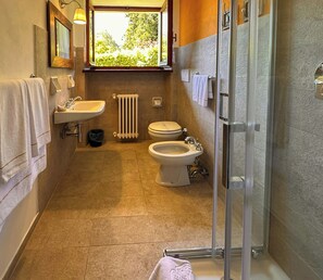 Un baño con ducha, dos inodoros, un lavabo y una ventana con vista a los árboles.