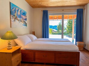 A bedroom with a bed, a nightstand with a lamp, a painting on the wall, and a view through the window.