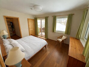 Ein Schlafzimmer mit einem Bett, einem Sessel, einem Holztisch, einer Lampe, einem Kleiderschrank und einem Fenster mit Vorhängen.