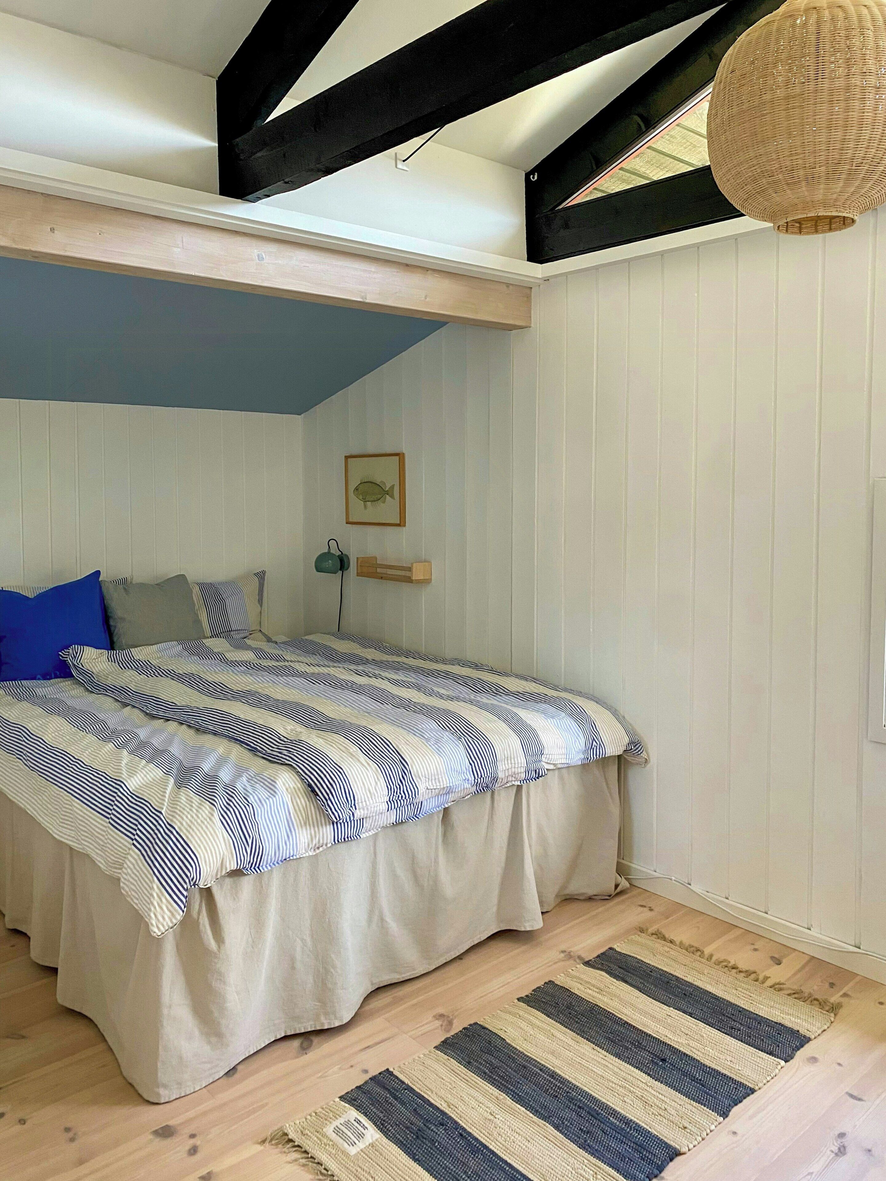 Ein Schlafzimmer mit Bett, blauer Wand, Holzbalke an der Decke, Pendelleuchte und einem gerahmten Bild an der Wand.
