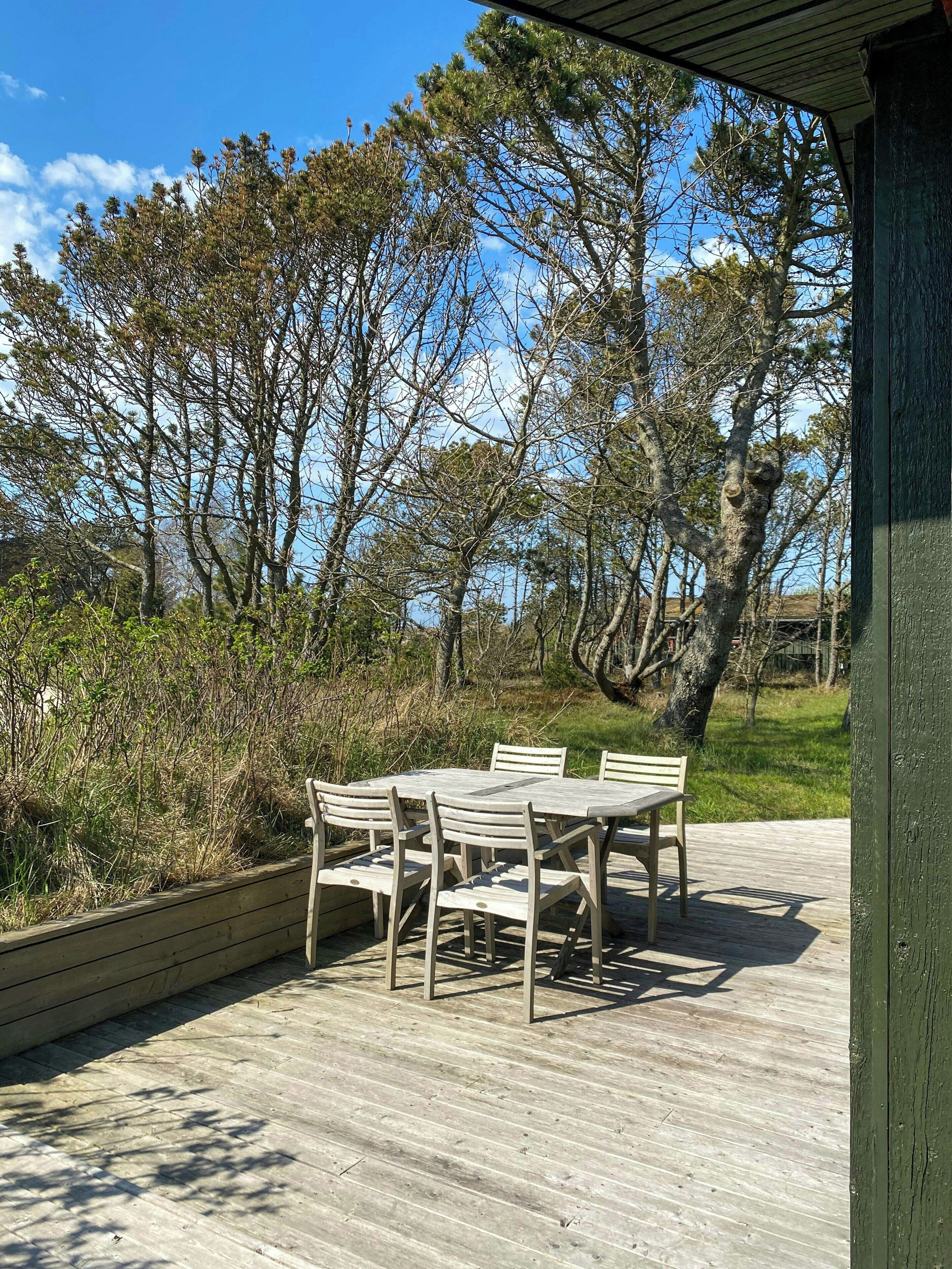 A wooden deck with a table and chairs, surrounded by trees and a clear sky.