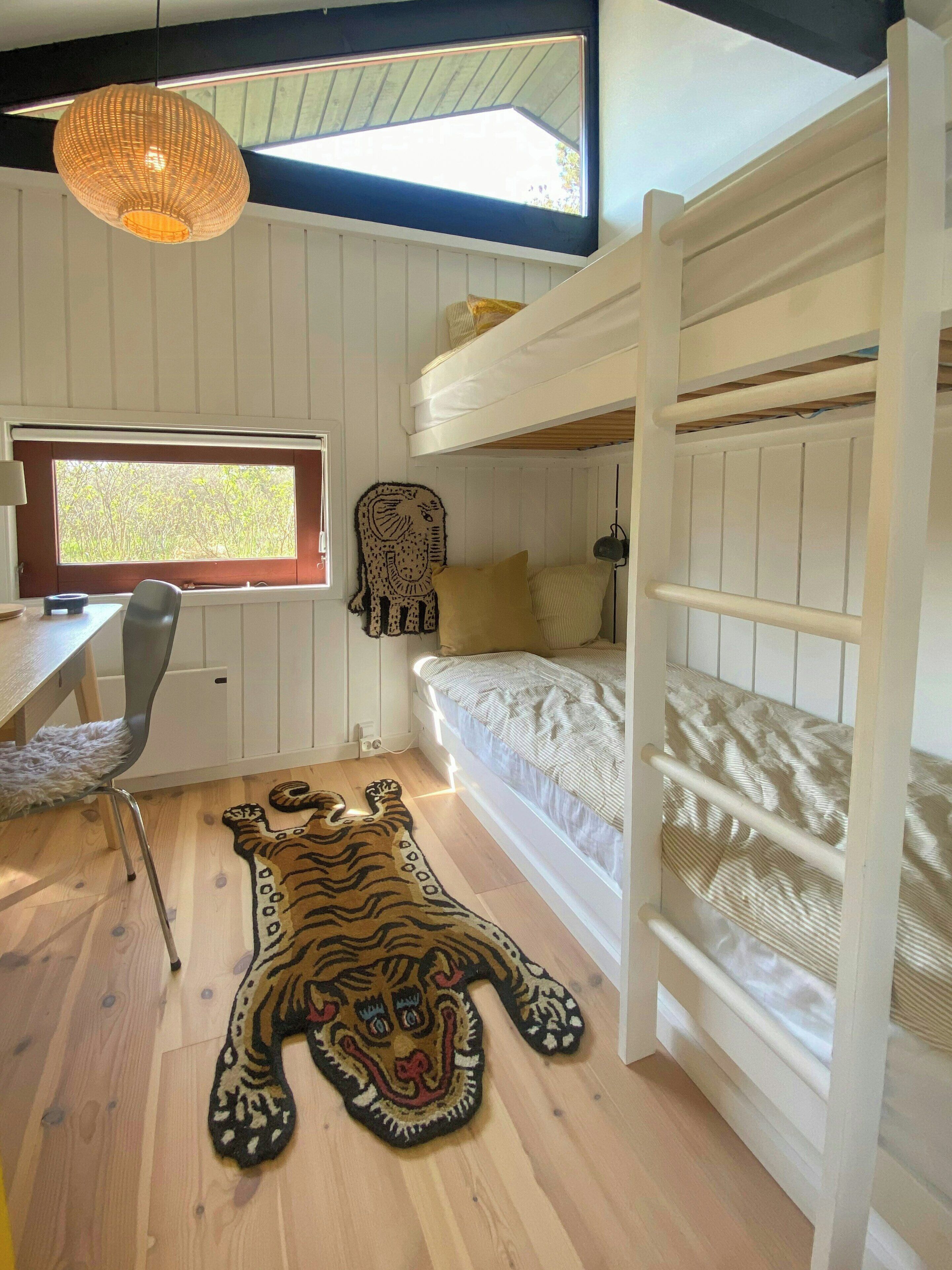 Une chambre avec un lit superposé, un bureau avec une chaise, une fenêtre et un tapis à motifs de tigre sur le sol.