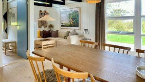 A dining area with a wooden table and chairs, a hanging light fixture, and a view of greenery outside. - Summer house vibe at Kandestederne (Skagen)