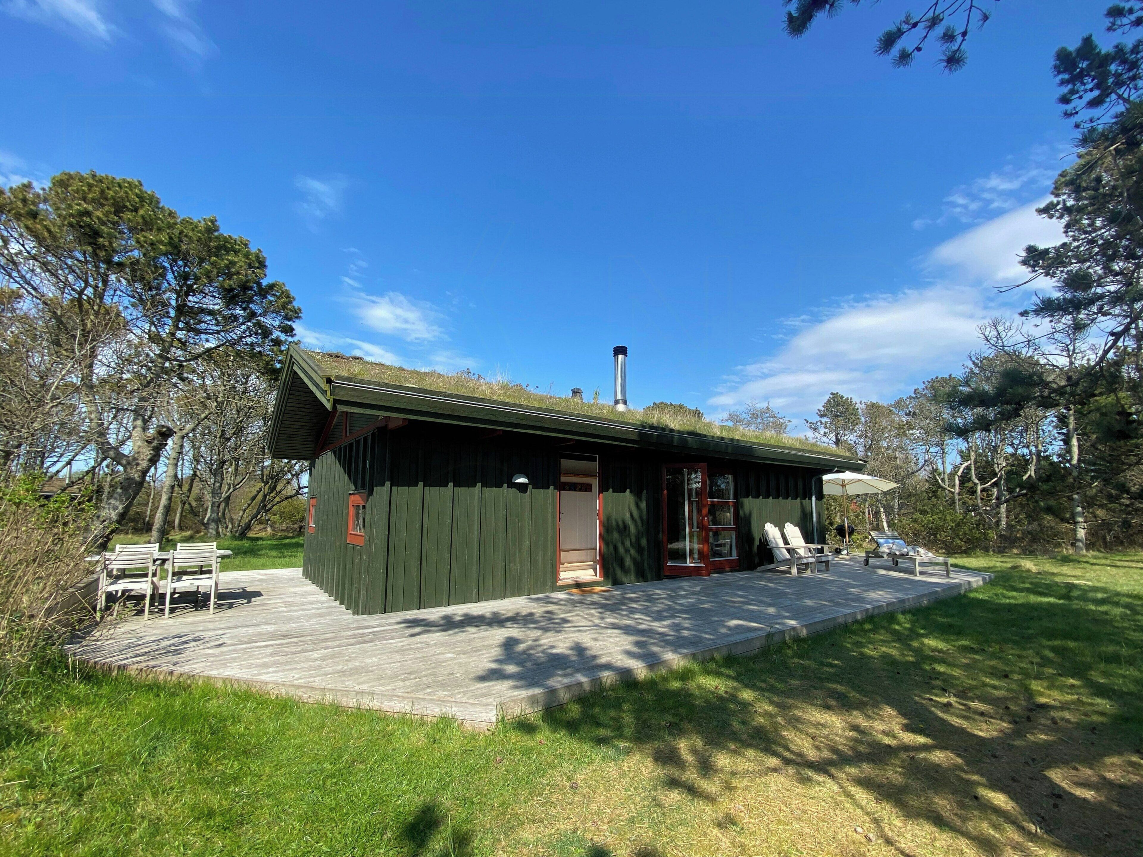 Una cabaña de madera con techo verde, chimenea y un patio con mobiliario exterior.