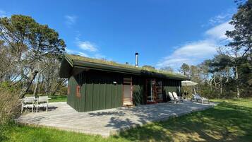 Una cabaña de madera con techo verde, chimenea y un patio con mobiliario exterior.