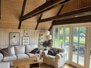 A cozy living room with a wooden ceiling, a beige sofa, a wooden coffee table, and a large window with a view of the outdoors.
