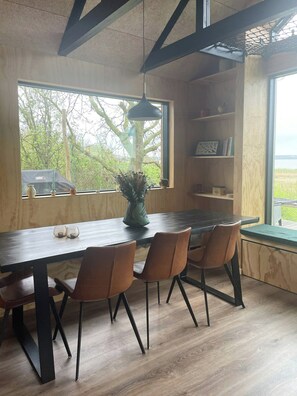 Un coin repas avec une table en bois, quatre chaises, un luminaire suspendu et une vue sur les arbres par une fenêtre.