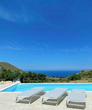 Ein Pool mit Liegestühlen, wolkenloser Himmel und Blick auf Meer und Hügel.
