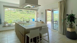 Una cuina moderna amb una illa gran, cadires blanques, vista a la vegetació i una porta corredissa de vidre.