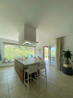 Eine moderne Küche mit einer großen Kochinsel, weißen Stühlen, Blick auf Grünflächen und einer Schiebeglastür.