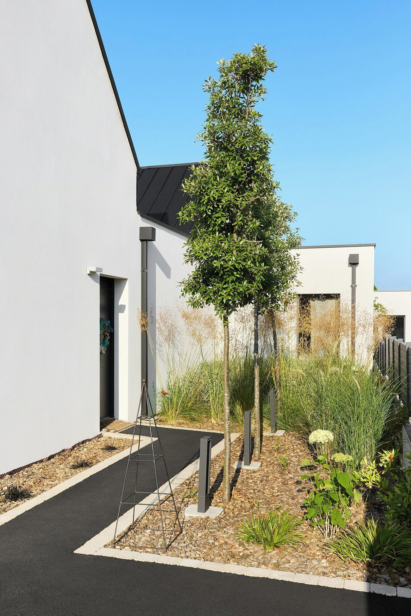 A modern building with a white exterior, a black roof, and a landscaped garden area with a tree and various plants.
