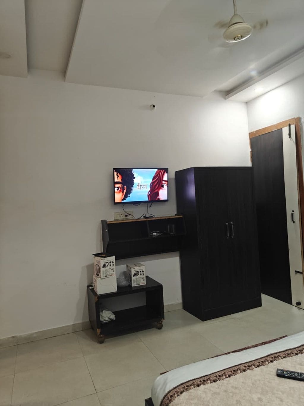 Un dormitorio con cama, televisor montado en la pared, armario y una mesita con cajas.
