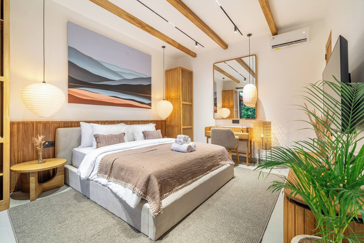 A modern hotel room with a large bed, a desk, a chair, a large mirror, and a potted plant.