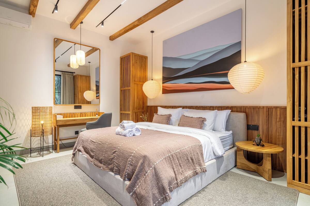 A modern bedroom with a large bed, wooden headboard, bedside tables, a mirror, and a painting on the wall.