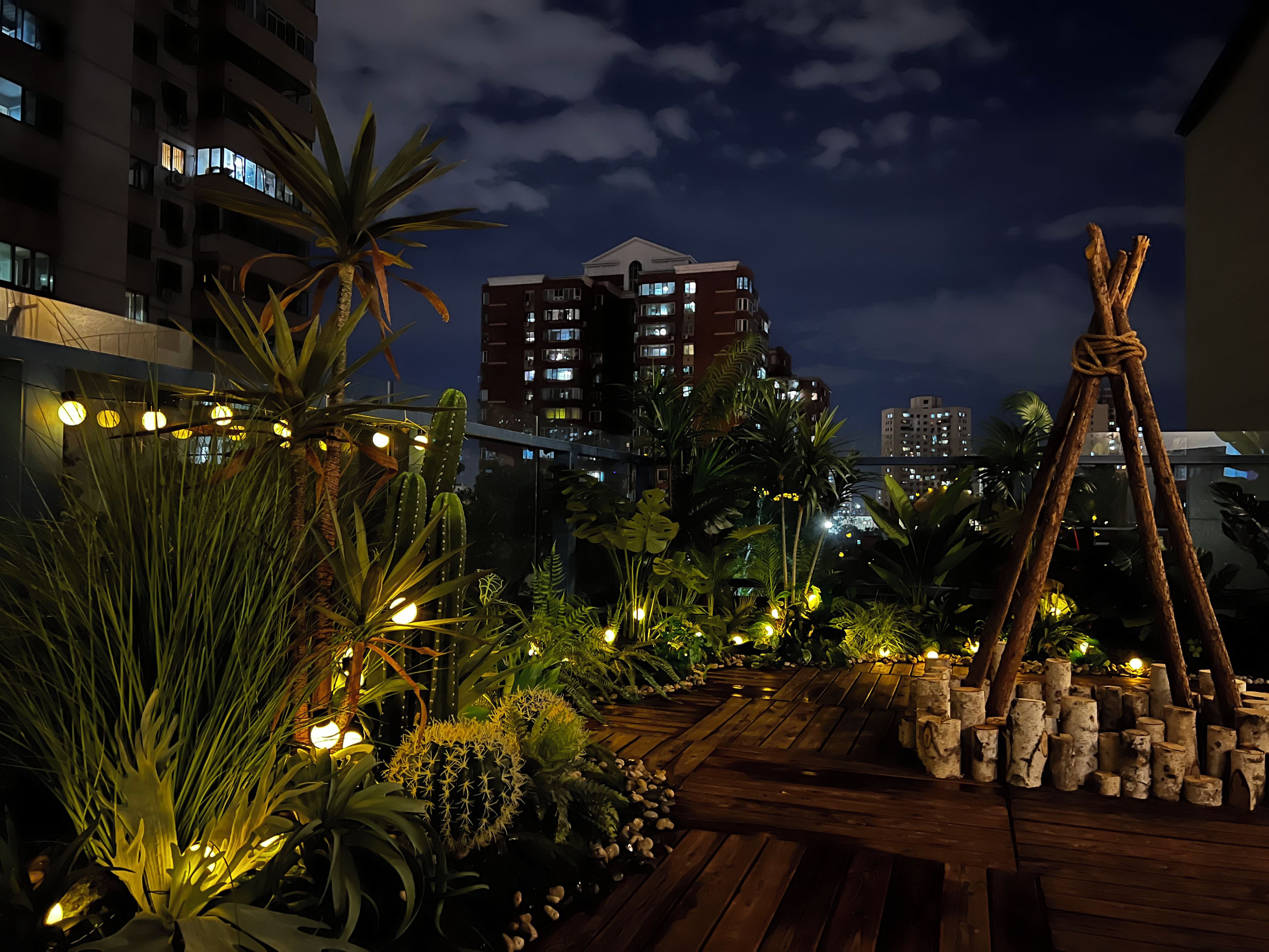 A rooftop garden with various plants and a wooden deck, surrounded by high-rise buildings.