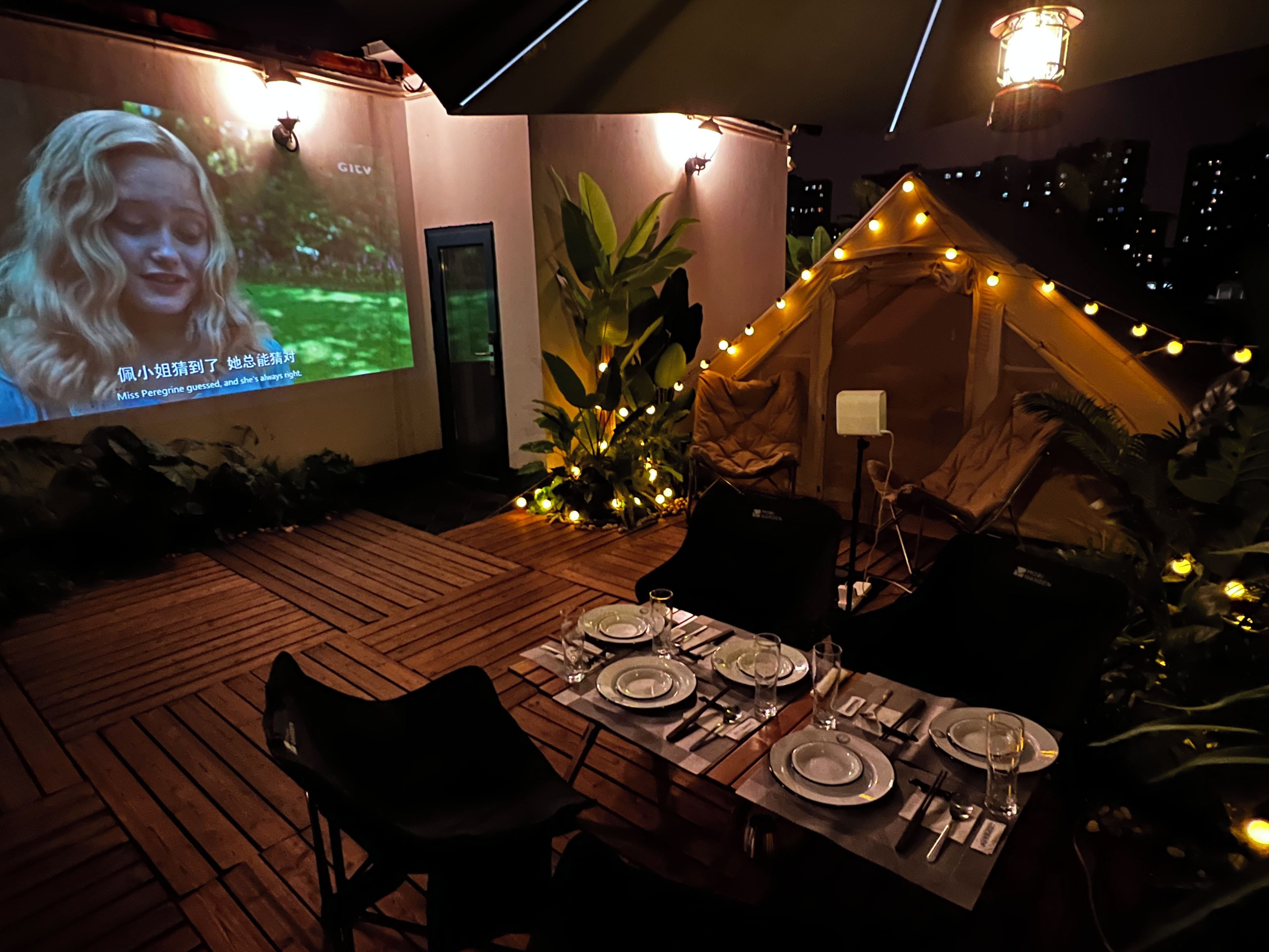 An outdoor dining area with a tent, string lights, and a projection screen displaying a movie.