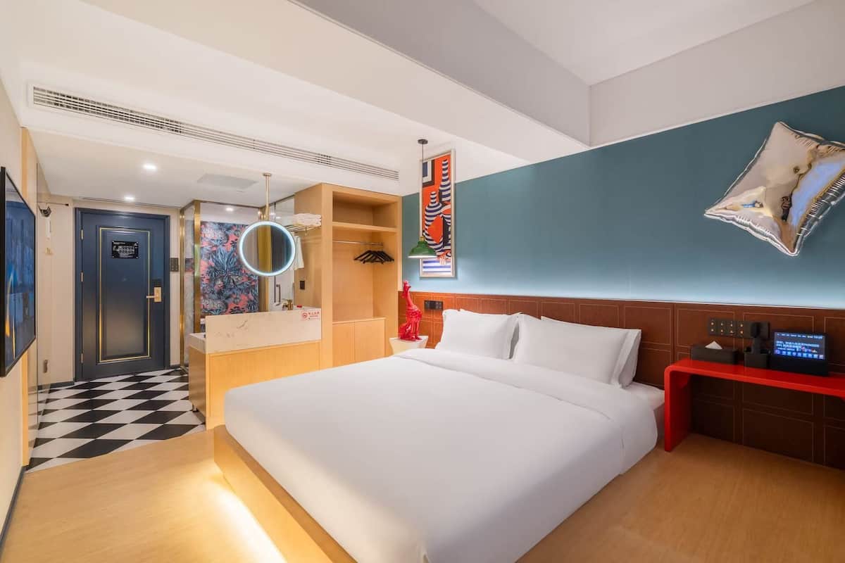Habitación de hotel con una cama grande, una mesita de noche roja, un espejo, un televisor montado en la pared y un baño con zona de ducha.