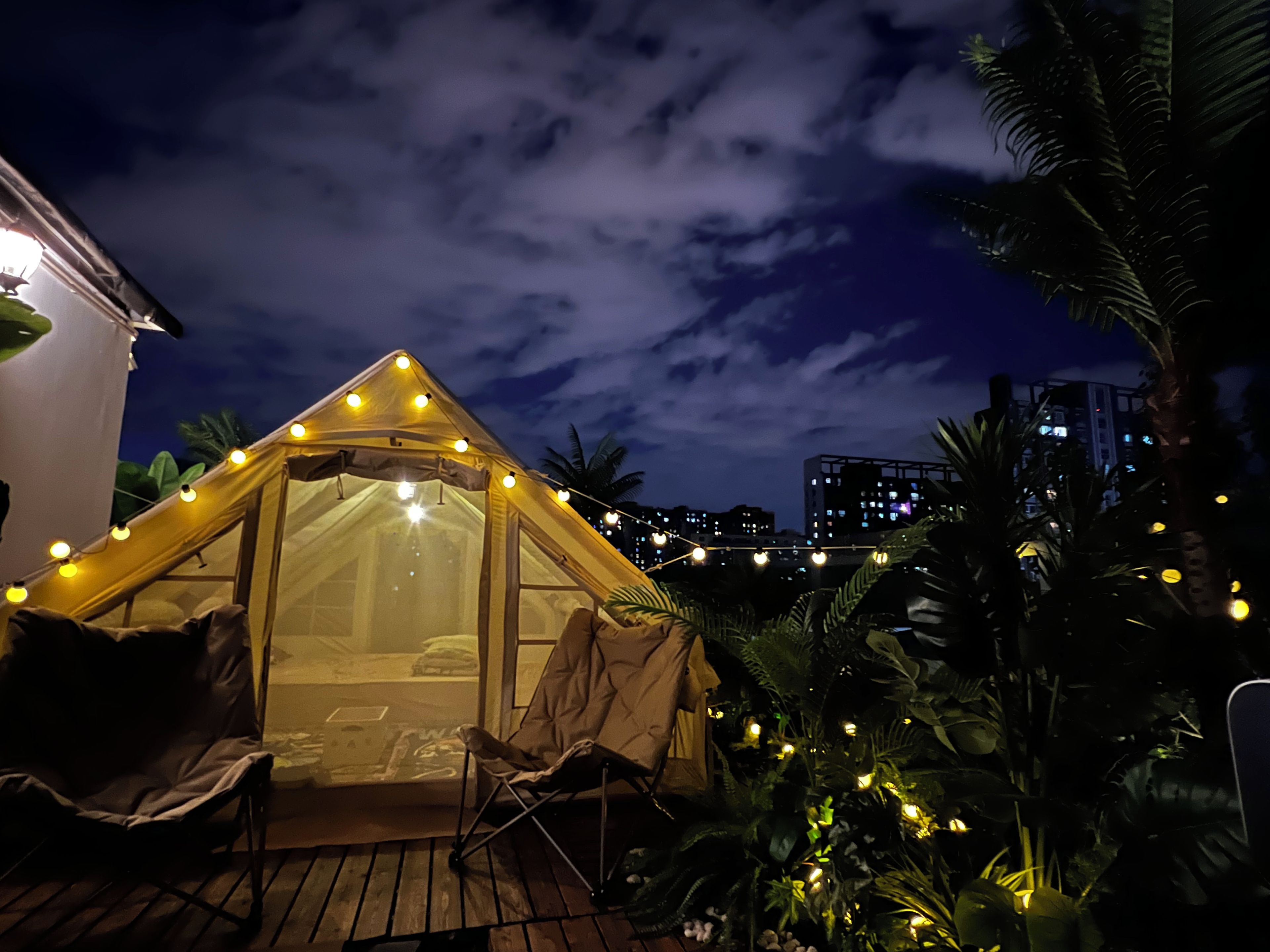 Une tente décorée de guirlandes lumineuses, une chaise et une table sur une terrasse en bois, avec des palmiers en arrière-plan.