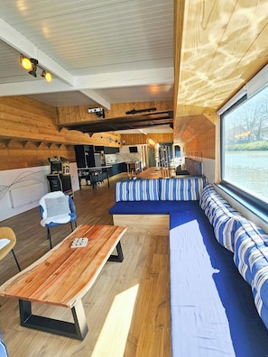 Interior de una cabaña con techo de madera, comedor y zona de estar con vista al agua.