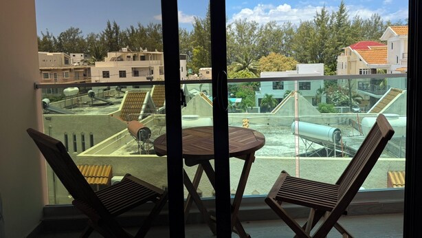 Un balcón con dos sillas de madera y una pequeña mesa, con vista a una terraza con edificios y árboles.