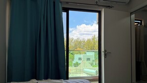 Un dormitorio con cama, ventana con cortinas, equipo de aire acondicionado y vista a edificios y árboles.