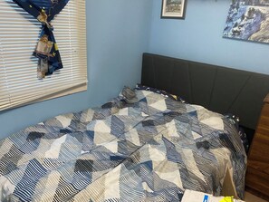 Une chambre à coucher avec un lit, une fenêtre avec des stores, un mur orné d’un tableau et une tête de lit.