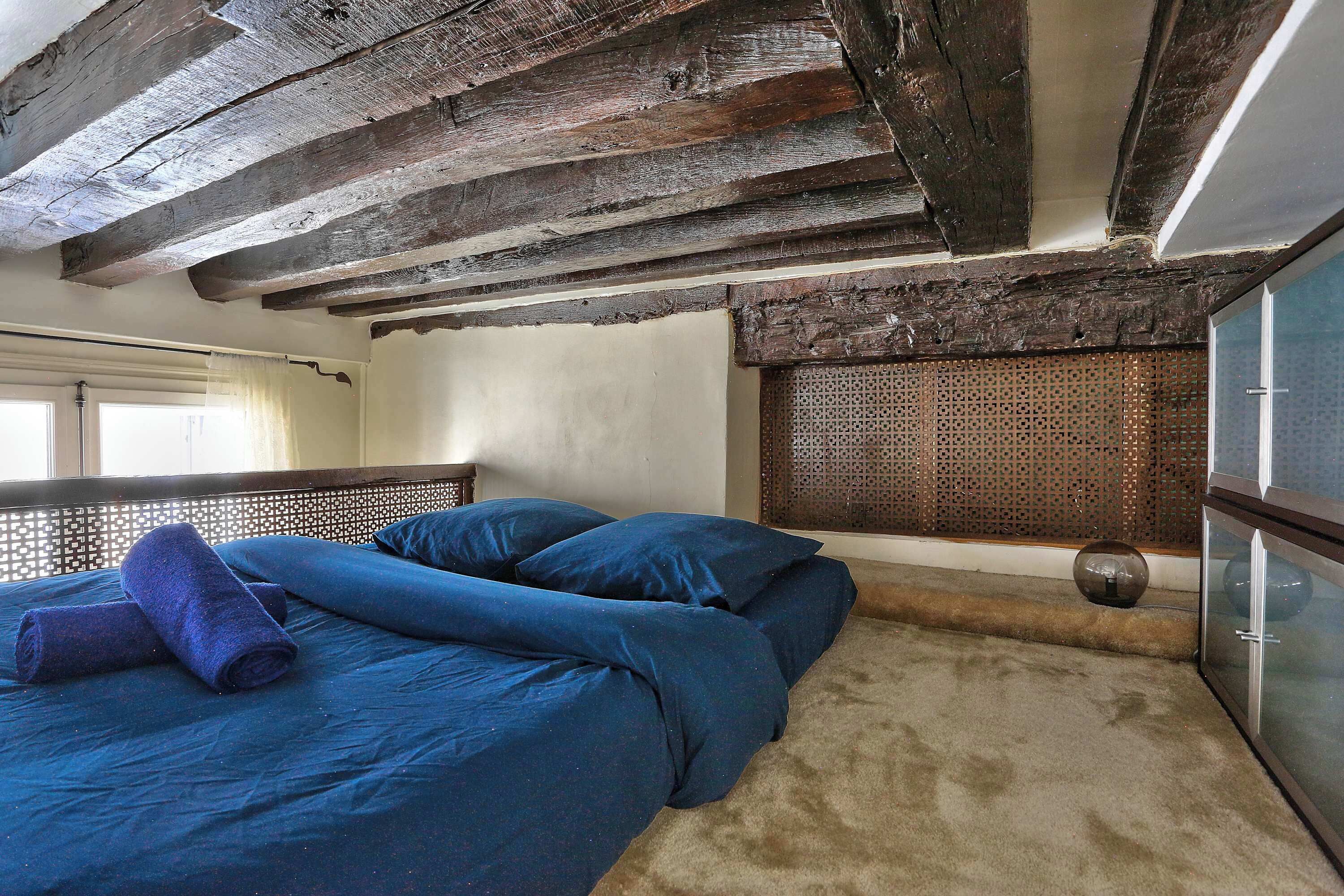 A room with a wooden ceiling, a bed with blue bedding, a window with a curtain, and a textured wall.