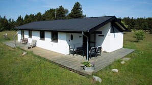 Et hvidt etplanshus med sort tag, træterrasse og et lille udendørs siddeområde.