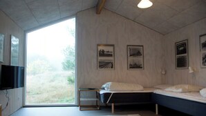 Et soveværelse med seng, et lille bord, et fjernsyn og et stort vindue med udsigt til grønne områder.
