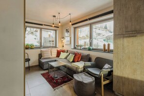 Ein modernes Wohnzimmer mit Sofa, Hocker und Couchtisch, großen Fenstern mit Blick auf Schnee und einer Holzkommode.