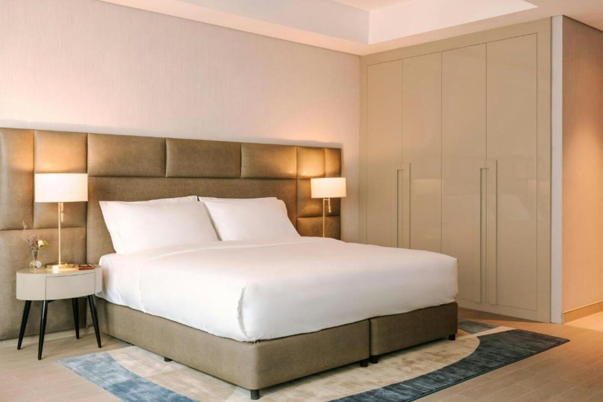 Quarto de hotel com uma cama grande, luminárias laterais, um guarda-roupa e um tapete.