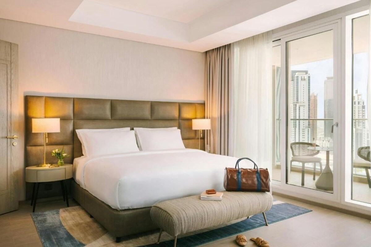 Habitación de hotel con una cama grande, lámparas de noche, un banco, un bolso y vistas de la ciudad a través de una puerta corrediza de vidrio.