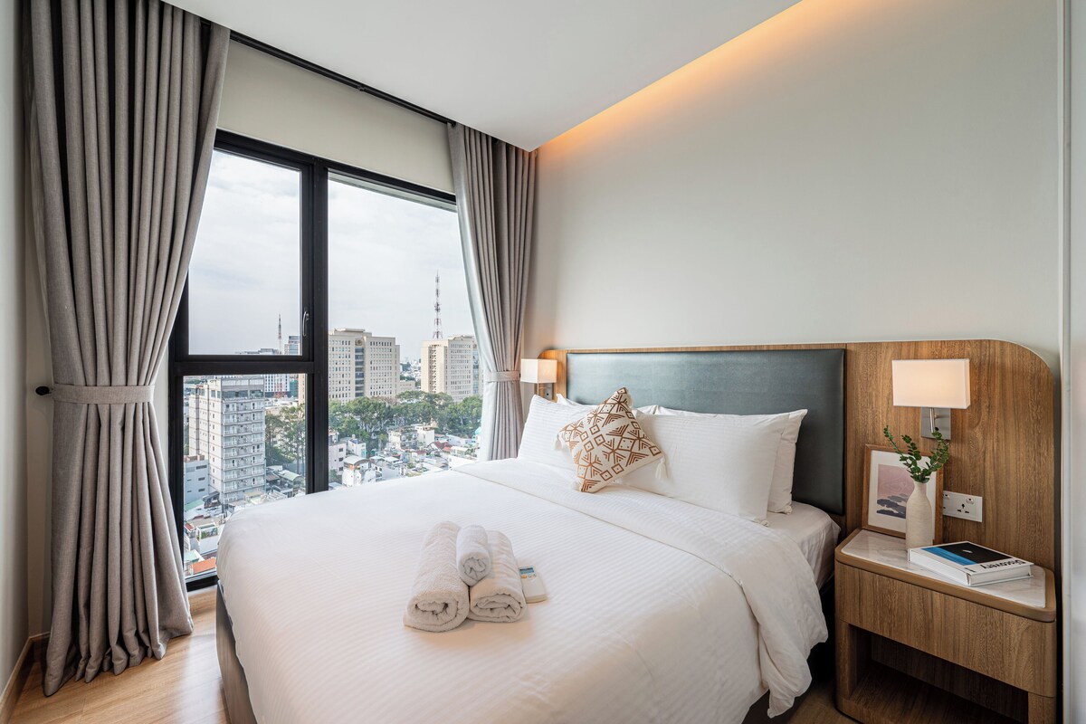 Ein Hotelzimmer mit einem großen Bett, einem Nachttisch mit Lampe und Blick auf die Stadt durch das Fenster.