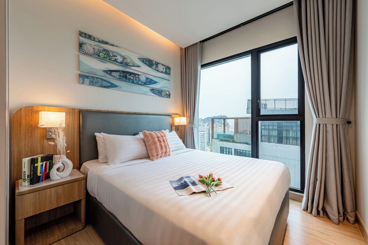 Ein modernes Hotelzimmer mit einem großen Bett, einem Nachttisch mit Lampe, einem Wandbild und Blick auf die Stadt durch ein großes Fenster.