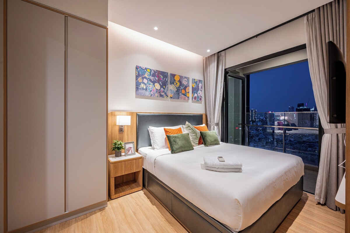 Ein modernes Hotelzimmer mit einem großen Bett, einem Nachttisch mit Lampe, Blick auf die Stadt und einem Kunstwerk an der Wand.