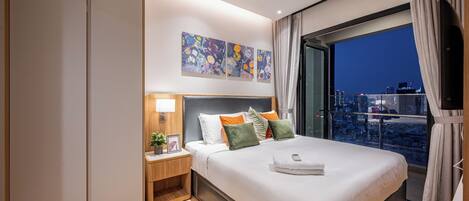 A modern hotel room with a large bed, a nightstand with a lamp, a city view, and artwork on the wall.