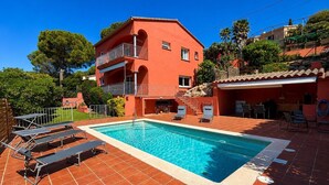 En röd villa med en swimmingpool, utomhusmöbler och en trädgård.