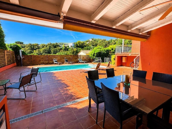 Ein Außenpoolbereich mit überdachter Terrasse, Liegestühlen und einem Esstisch.