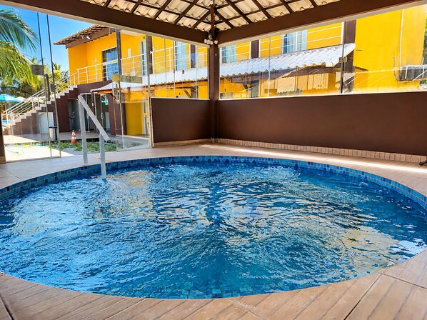 A swimming pool with a tiled edge, a covered area with a roof, and a building with yellow walls and a brown roof in the background. - Pousada X (Saquarema)