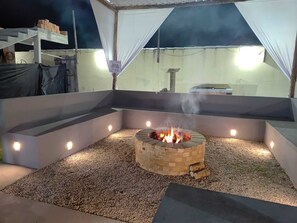 An outdoor fire pit area with a brick fire pit, surrounded by gravel, and illuminated by spotlights. - Pousada X (Saquarema)