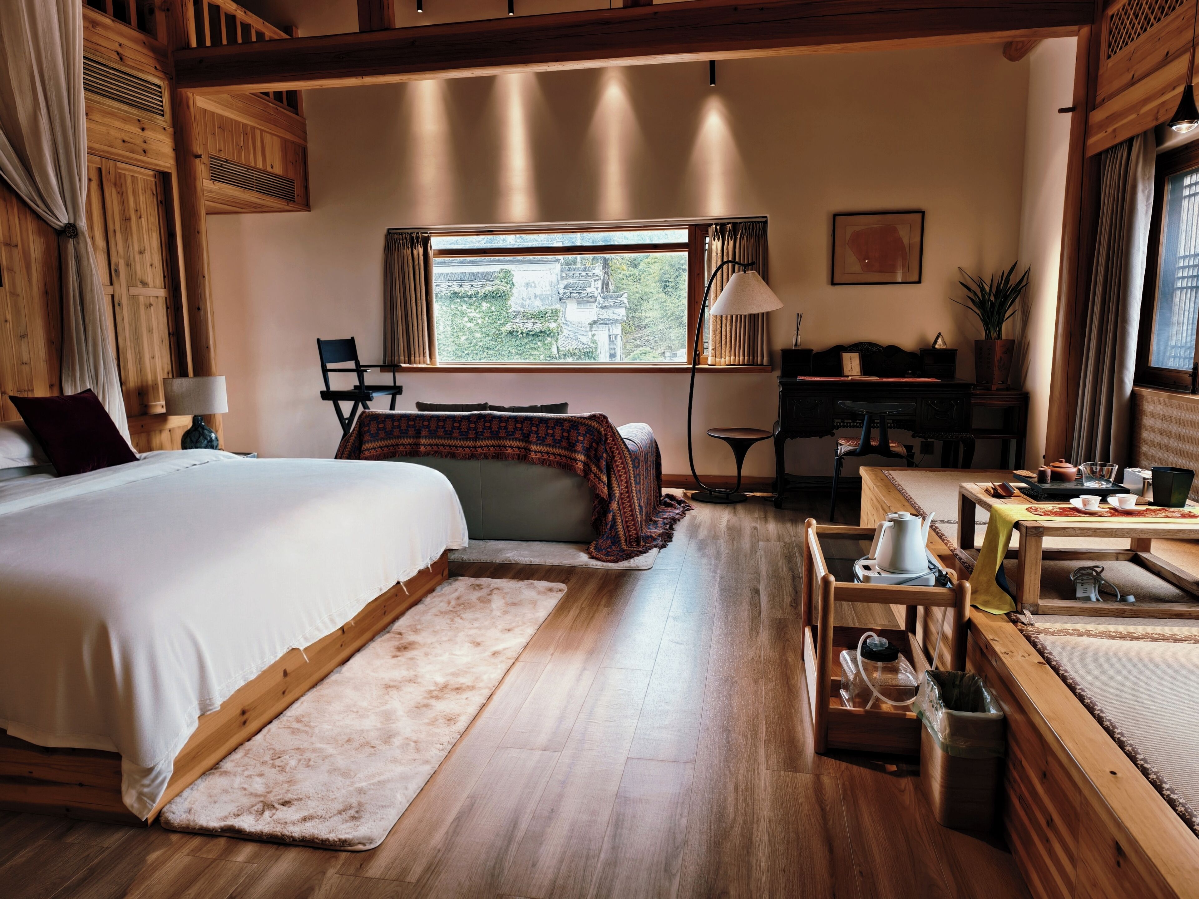 Una habitación con una cama grande, un escritorio con computadora, una silla, un ventanal con vistas y una mesita con tetera.