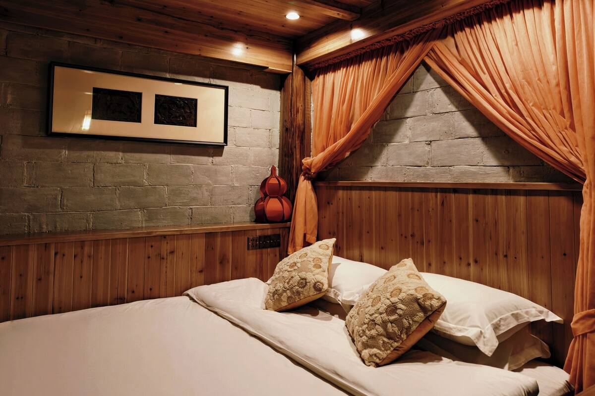 Habitación con paneles de madera, una cama, almohadas, un cuadro enmarcado y una ventana con cortinas.