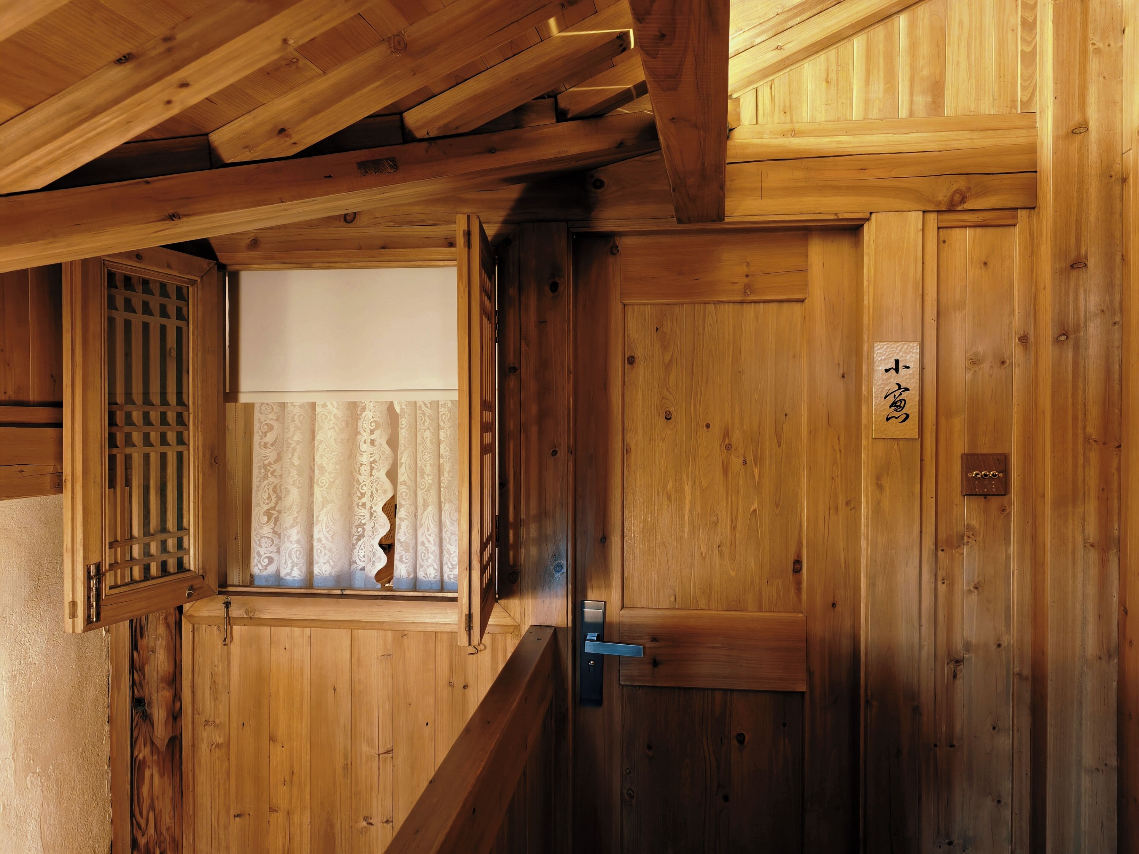 Un interior de madera con una puerta corrediza, una ventana con cortinas estampadas y un letrero de madera con caracteres japoneses.